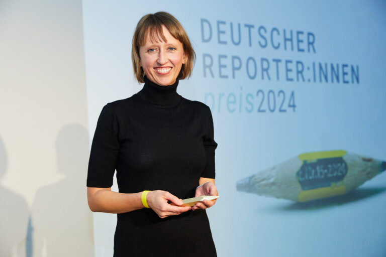 Eine Frau mit schulterlangen dunkelblonden Haaren in schwarz gekleidet vor einer Wand mit der Aufschrift: Deutscher Reporter:innenpreis 2024