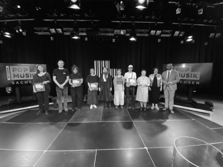 Eine Gruppe Menschen auf einer Bühne, vier von ihnen halten Urkunden in der Hand. Im Hintergrund die Schrift: Wettbewerb Popmusik Sachsen.