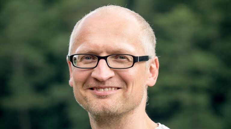 Lächelnder weißer Mann mit gräulicher Halbglatze und schwarzer Brille, im Hintergrund verschwommene Hecke